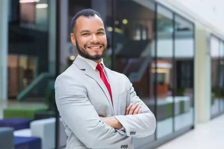 Cheerful businessman smiling at camera. Portrait of confident African American businessman in suit standing with crossed arms outside office building and looking at camera. Business concept